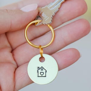 May include: A set of keys with a gold-colored key ring and a round, light green keychain. The keychain has a black outline of a house with a heart inside. The keys are held in a hand with painted nails.