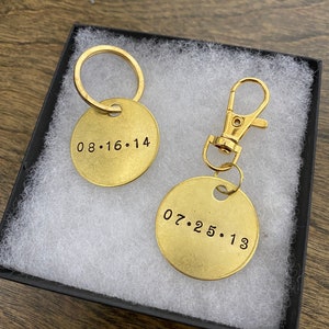 May include: Two gold keychains with round metal tags. The tag on the left keychain has the date "08.16.14" engraved on it. The tag on the right keychain has the date "07.25.13" engraved on it.