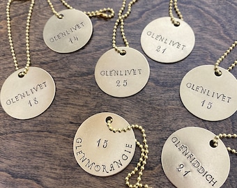 May include: Seven circular brass tags with gold-toned ball chains. Each tag is stamped with black text including brand names like Glenlivet, Glenmorangie, and Glenfiddich, along with specific age numbers such as 14, 15, 18, 21, and 25.