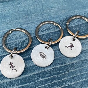 May include: Three silver keychains with round metal tags. Each tag features a different animal silhouette: a lizard, a chameleon, and a frog.