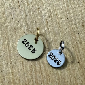 May include: Two small, round metal charms with the year "2025" stamped on them. One charm is gold-toned with a gold-colored loop, and the other is silver-toned with a silver loop. The charms are on a wooden surface.