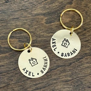 May include: Two round gold-toned metal keychains, each featuring a stamped house icon with a heart in the center and the names AXEL and SARAHI engraved in black lettering. Each circular tag is attached to a matching gold-toned metal split ring.