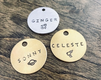 May include: Three round metal pet tags with engraved names and space-themed designs. The tags are gold and silver in color. The names are "Ginger", "Sonny", and "Celeste".