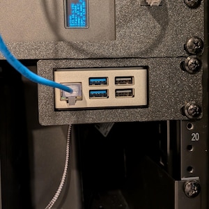 May include: A black PiKVM V4 plus server rack mount unit featuring a small blue digital display, a silver front panel with four USB ports, and an Ethernet port connected to a blue cable. The device is secured with black screws in a rack.