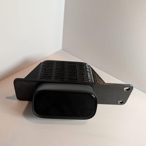 Puede incluir: Un dispositivo rectangular negro con una parte delantera curvada, montado en un soporte negro con una parte superior ranurada y dos agujeros. El dispositivo está sobre un fondo blanco.