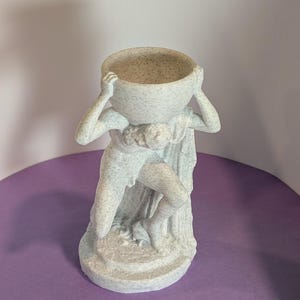 May include: A light gray statue of a figure holding a large bowl above its head. The figure is in a classical pose, with a draped cloth and detailed musculature. The bowl and figure have a speckled texture, and the statue is on a round base.