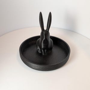 Puede incluir: Porta anillos de cerámica negra con forma de conejo. El conejo está sentado en un plato de cerámica negra.