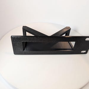 Puede incluir: Un soporte negro rectangular con una abertura central y dos orificios de montaje. El soporte tiene una estructura de soporte en ángulo. El artículo es probablemente para montar o soportar equipos.