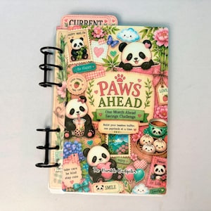 May include: A spiral-bound savings challenge planner titled Paws Ahead One Month Ahead Savings Challenge. The cover features illustrations of pandas, bamboo, flowers, and food items with text including The Humble Budgeter and Current.