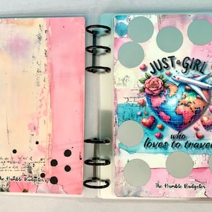 May include: A clear binder with a pink and cream abstract design on the left and a travel-themed design on the right. The right side features a globe, airplane, hearts, and the text "JUST a GIRL who loves to travel."