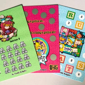 May include: Three rectangular planner inserts featuring Rugrats characters. The green insert has a block tracking grid. The pink insert has circular scratch-off spots and the text A baby's gotta do. The blue insert features alphabet blocks and the text Rugrats.