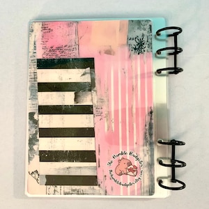 May include: A planner cover with a collage design featuring pink, black, and white elements. The design includes stripes, text, and a logo with a pig holding a heart. The planner has black metal rings.