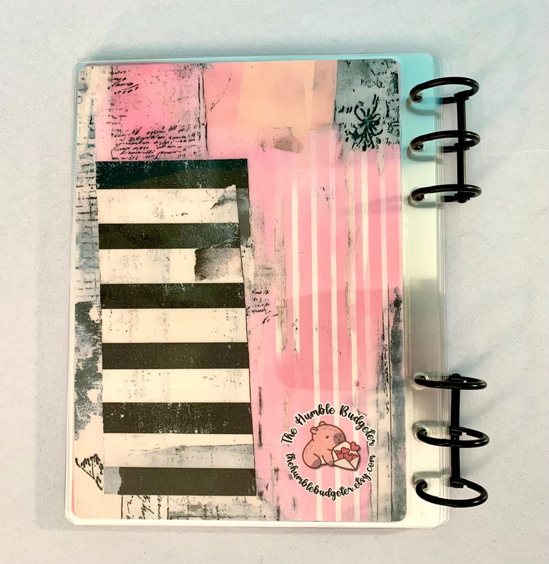 May include: A planner cover with a collage design featuring pink, black, and white elements. The design includes stripes, text, and a logo with a pig holding a heart. The planner has black metal rings.