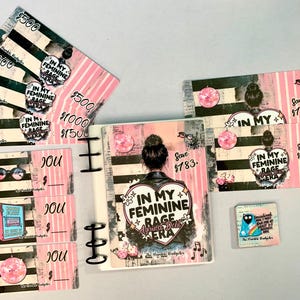 May include: A collection of pink and black budget-themed accessories. Includes cash envelopes, a binder, and stickers. The items feature text and graphics, including the phrase "In My Feminine Rage Era."