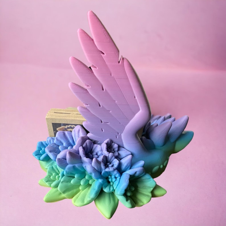 May include: A 3D printed bird figurine with outstretched wings, painted in a gradient of pink and purple. The bird is perched on a bed of colorful flowers, with shades of blue, green, and purple.