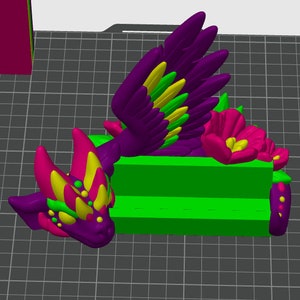 May include: A 3D printed figurine of a purple and yellow dragon with green accents. The dragon is lying down with its wings spread out. The dragon is on a green platform with a pink flower.