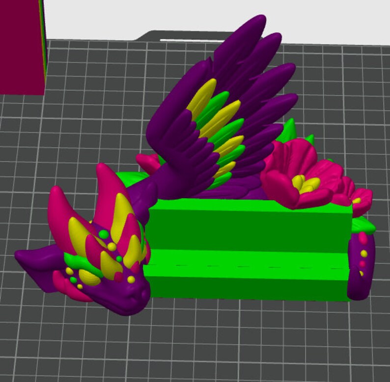 May include: A 3D printed figurine of a purple and yellow dragon with green accents. The dragon is lying down with its wings spread out. The dragon is on a green platform with a pink flower.