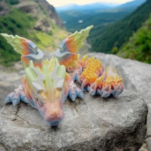 Può includere: Una statuetta di drago colorata in più pezzi, con ali, coda e testa. Il drago è fatto di un materiale liscio di colore pastello e ha un aspetto fantasioso e da cartone animato.