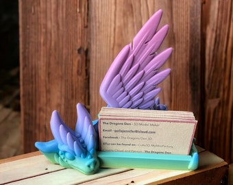 May include: A 3D printed dragon-shaped business card holder with a purple and blue gradient. The holder is made of plastic and has a small dragon head with wings. The card holder is sitting on a wooden surface. The Dragons Den - 3D Model Maker. Email: gollajennifer@icloud.com. Facebook: The Dragons Den 3D. STLs can be found on - Cults 3D, MyMiniFactory, Creality Cloud, and Patreon The Dragons Den.