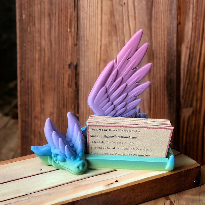 May include: A 3D printed dragon-shaped business card holder with a purple and blue gradient. The holder is made of plastic and has a small dragon head with wings. The card holder is sitting on a wooden surface. The Dragons Den - 3D Model Maker. Email: gollajennifer@icloud.com. Facebook: The Dragons Den 3D. STLs can be found on - Cults 3D, MyMiniFactory, Creality Cloud, and Patreon The Dragons Den.