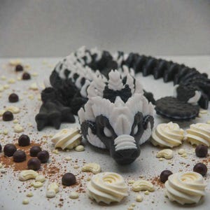 May include: A detailed, 3D-printed badger figurine in black and white, with a long, segmented body. The badger is surrounded by small, round chocolate candies, white frosting swirls, and cocoa powder, creating a whimsical, dessert-themed display.