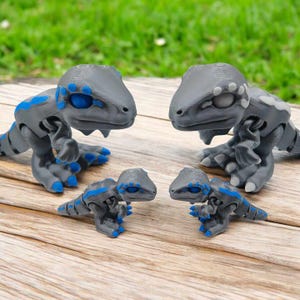 May include: Four gray dinosaur figurines with blue accents. The two larger dinosaurs are standing upright, while the two smaller dinosaurs are lying down. The dinosaurs have a textured surface and are made of plastic.