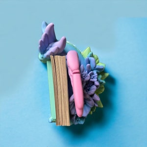 May include: A pastel blue and green floral bookend with a pink bookmark. The bookend is shaped like a flower with a book resting on top.