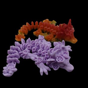 May include: Two small, detailed dragon figurines. One is a vibrant red and orange, the other is a soft purple. Both have floral details and are made of resin.
