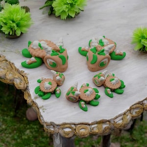 Könnte beinhalten: Fünf handgefertigte Keramikfiguren einer mythischen Kreatur in Grün, Braun und Weiß. Die Figuren sind auf einer weißen Oberfläche mit dekorativem goldenen Rand angeordnet.