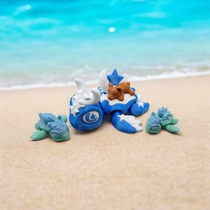May include: Three blue and white sea turtle figurines on a sandy beach. The largest turtle has a brown and white star design on its shell and a crescent moon on its head. The two smaller turtles are green and blue.