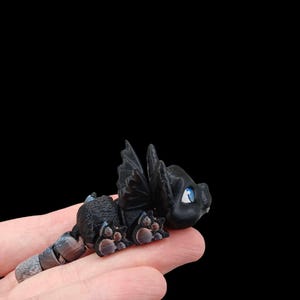 May include: A small, black, 3D printed dragon figurine with blue eyes. The dragon is posed in a crawling position with its wings spread out.