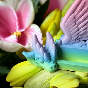May include: A rainbow-colored dragon figurine with wings, sitting on a bed of green and yellow flowers. The dragon has a spiky head and a long tail.