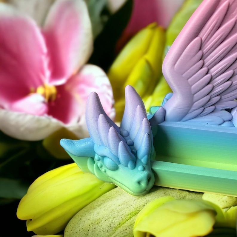 May include: A rainbow-colored dragon figurine with wings, sitting on a bed of green and yellow flowers. The dragon has a spiky head and a long tail.