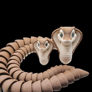 Op de afbeelding: Twee bruine en witte cobra-slangenfiguren, een groter dan de andere, met blauwe ogen. De grotere cobra staat rechtop met zijn kap open. De kleinere cobra is opgerold met zijn kop omhoog. Beide figuren zijn gemaakt van een glad, getextureerd materiaal.