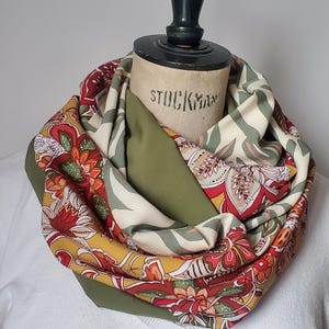 Pode incluir: Uma echarpe infinita com padrão verde, creme e vermelho com um design floral. A echarpe está dobrada e exibida em um manequim.