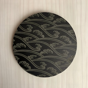 May include: A round, black coaster with a wave pattern in gray. The design features stylized waves, creating a repeating pattern across the surface. The coaster is made of a solid material.