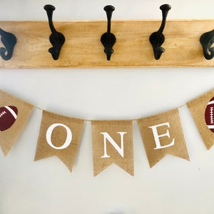 May include: A burlap banner with the word "ONE" in white letters and two brown footballs on either end. The banner is strung on a white wall.