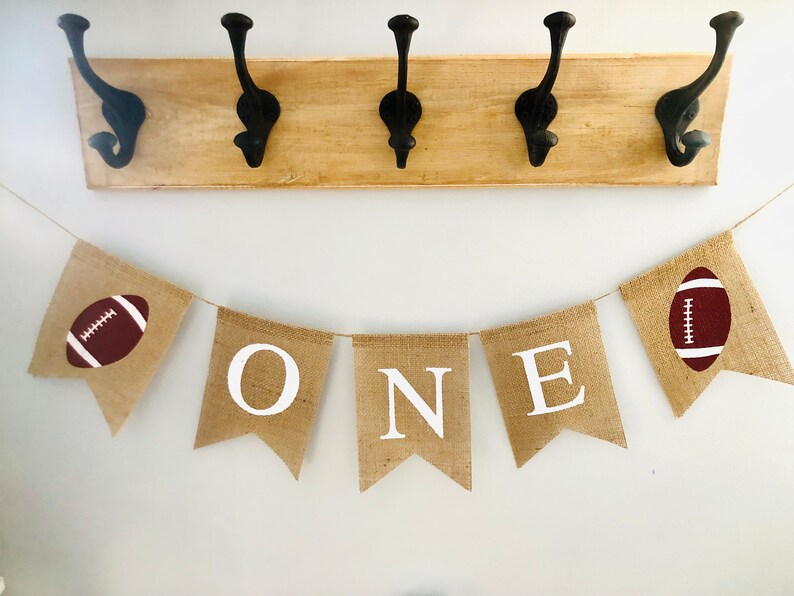 May include: A burlap banner with the word "ONE" in white letters and two brown footballs on either end. The banner is strung on a white wall.