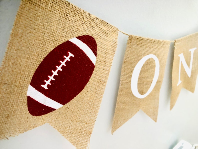 May include: A burlap banner with the word "ONE" in white letters and a red and white football on one of the flags.