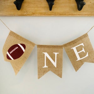 May include: A burlap banner with the word "ONE" in white letters and a brown football on the first flag. The banner is strung on a white wall.