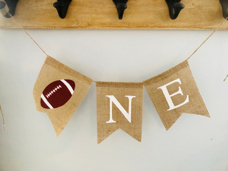 May include: A burlap banner with the word "ONE" in white letters and a brown football on the first flag. The banner is strung on a white wall.