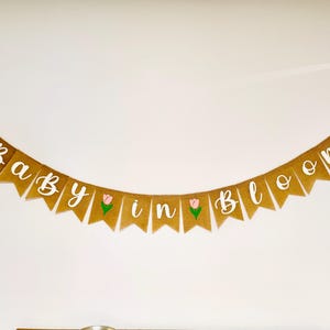 May include: A burlap banner with the text "Baby in Bloom" in white lettering. The banner has pink and green flower accents.