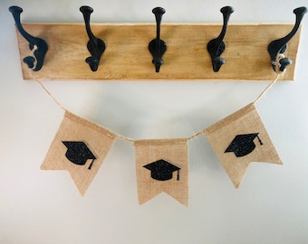 May include: A graduation banner with three burlap flags, each adorned with a black graduation cap. The flags are strung on twine and hang below a wooden board with black metal hooks, creating a festive decoration.