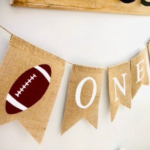 May include: A burlap banner with the word "ONE" in white letters and a brown football on each end. The banner is strung with twine.