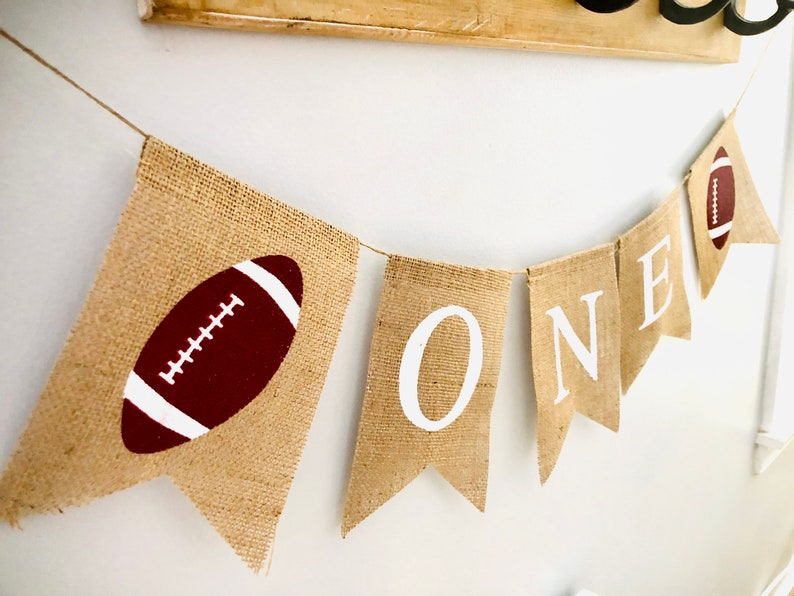 May include: A burlap banner with the word "ONE" in white letters and a brown football on each end. The banner is strung with twine.