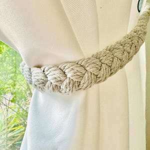 May include: A beige rope curtain tieback, braided in a thick, decorative style. The tieback is looped around a white curtain.