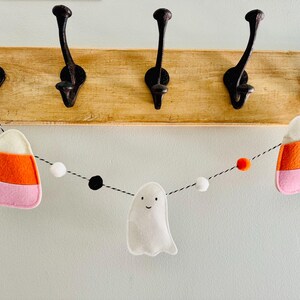 May include: A Halloween-themed garland featuring felt candy corn, a smiling ghost, and colorful pom-poms. The garland is strung on a black and white string and is hanging below a wooden board with black hooks.