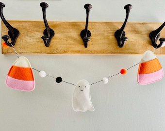 May include: A Halloween-themed garland featuring felt candy corn, a smiling ghost, and colorful pom-poms. The garland is strung on a black and white string and is hanging below a wooden board with black hooks.