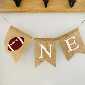 May include: A burlap banner with the word "ONE" spelled out in white letters. The first "O" is replaced with a brown and white football.
