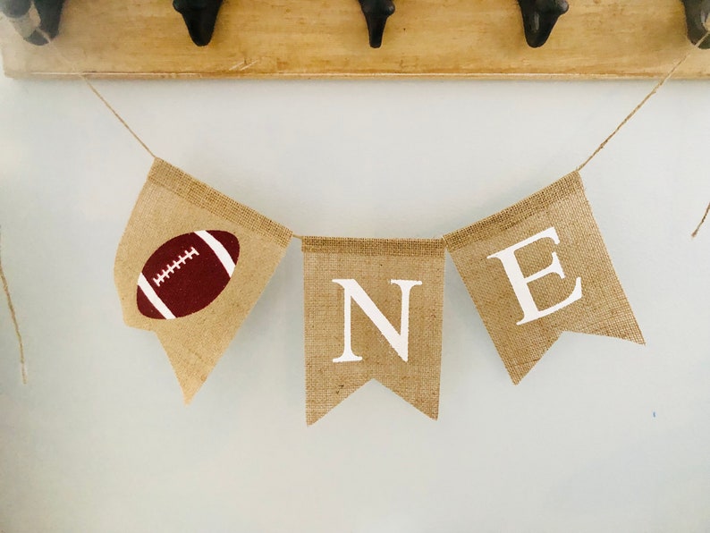 May include: A burlap banner with the word "ONE" spelled out in white letters. The first "O" is replaced with a brown and white football.